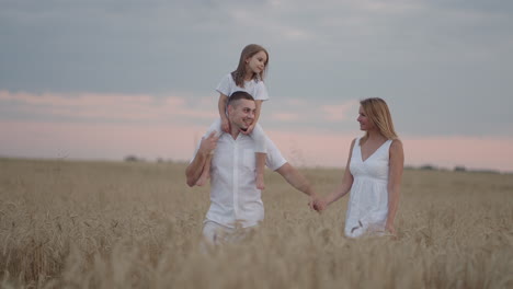 Young-couple-of-parents-with-girl-children-holding-hands-of-each-other-and-running-through-wheat-field-at-sunset.-Happy-family-jogging-among-barley-meadow-and-enjoying-nature-together.-Slow-motion